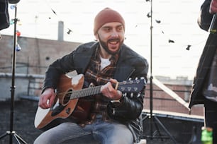 Young bearded man. Guy with acoustic guitar sings. Friends have fun at rooftop party with decorative colored light bulbs.