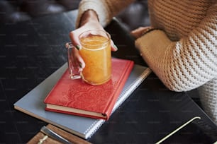 Close up foto da bebida laranja que segura a mão da mulher e fica no livro com capa vermelha.