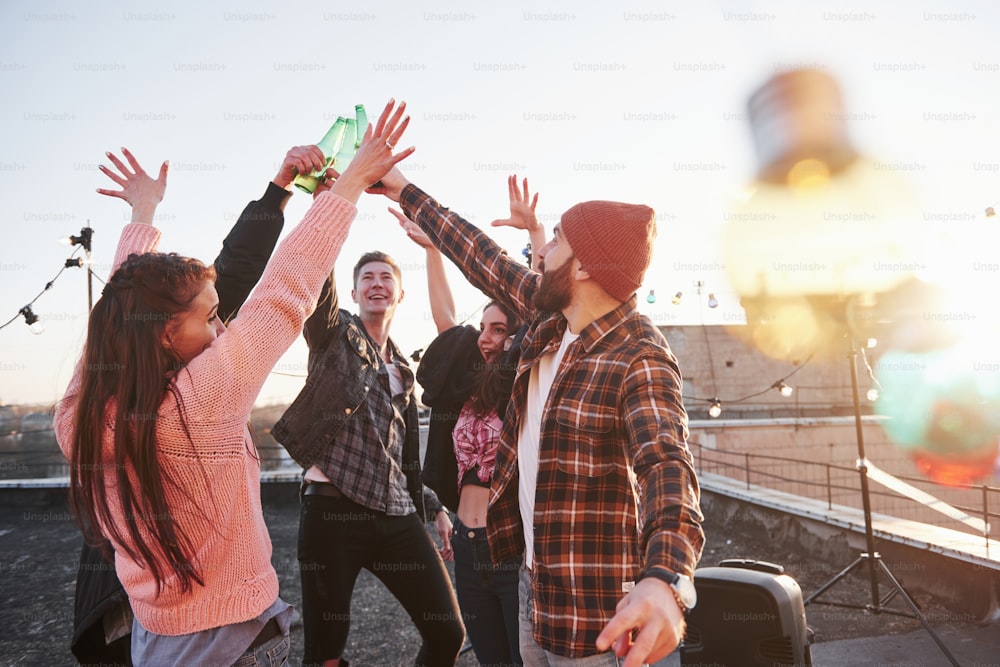 Decorative festive light bulbs. Holidays on the rooftop. Cheerful group of friends raised their hands up with alcohol.