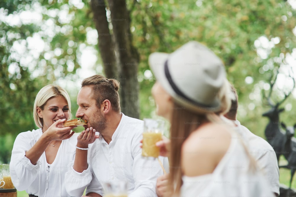 Woman feeds her husband with delicious sandwich outdoor with friends at daytime.
