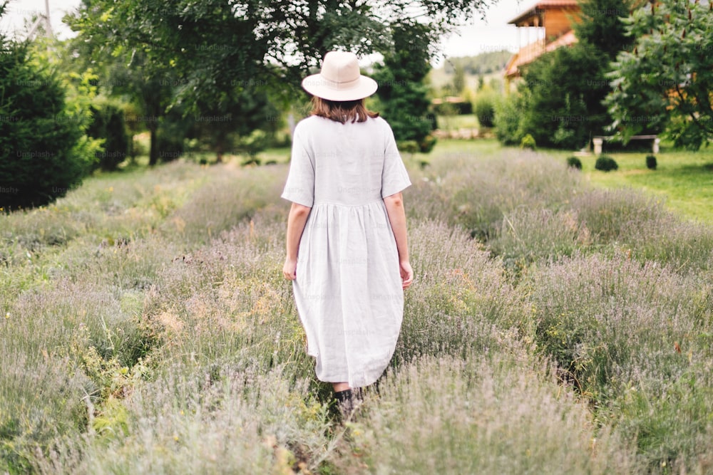 Stylish hipster girl in linen dress and hat walking in lavender field and relaxing. Happy bohemian woman enjoying lavender aroma. Back view. Atmospheric calm rural moment. Space for text