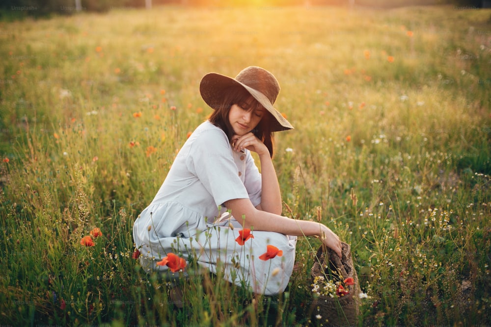 Stylish girl in linen dress sitting in poppy meadow in sunset light with flowers in rustic straw basket. Boho woman  in hat relaxing in summer field. Atmospheric moment, space text