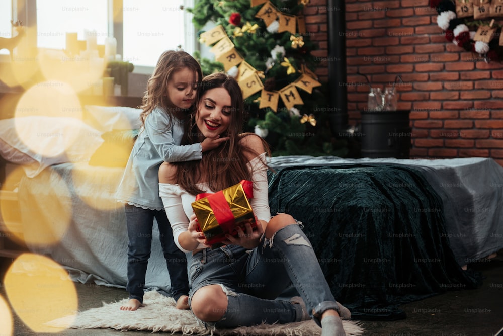 Look what I have for you. Mother and daughter sits in holiday decorated room and holds gift box.