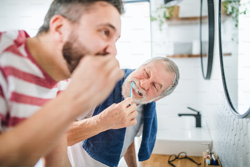 An adult hipster son and senior father brushing teeth in the bathroom indoors at home.