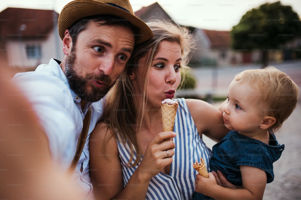 夏に屋外でアイスクリームを食べる両親と小さな幼児の女の子、自撮り写真を撮るときに顔をしかめます。