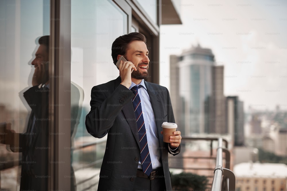 Take a pause. Waist up portrait of cheerful businessman talking by phone while standing on office balcony with cup of coffee. Copy space on right
