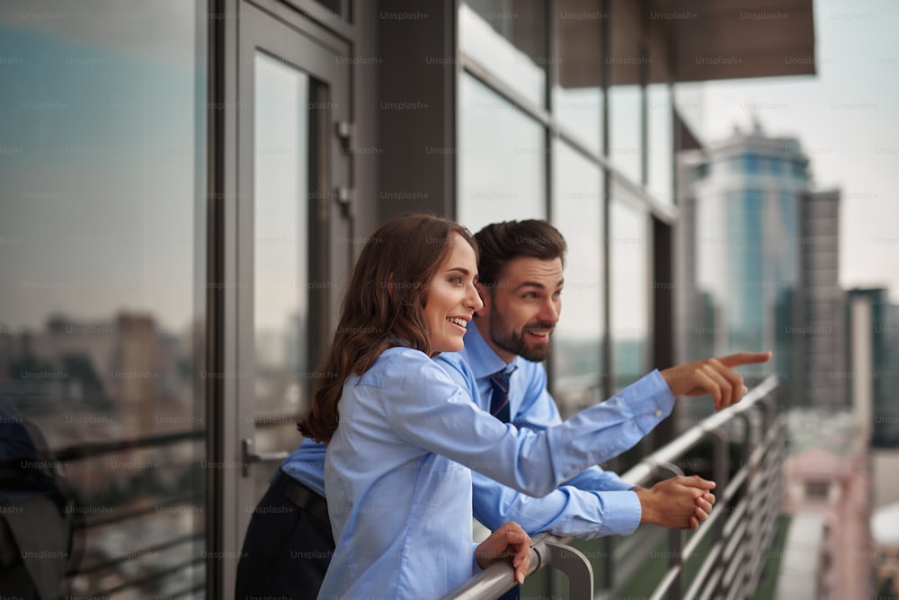 Concept of friendly communication between coworkers. Waist up portrait of smiling woman showing something to man from office balcony while have work break