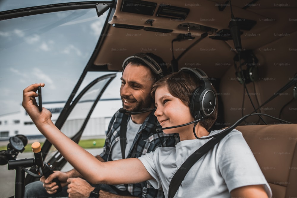 Side view smiling child and positive father taking photo on phone while wearing headphones. They locating in cabin of modern helicopter
