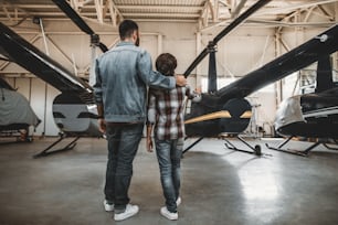 Full length kid pointing hand on big rotor plane. Man hugging him while listening his description of vehicle. They turning back to camera