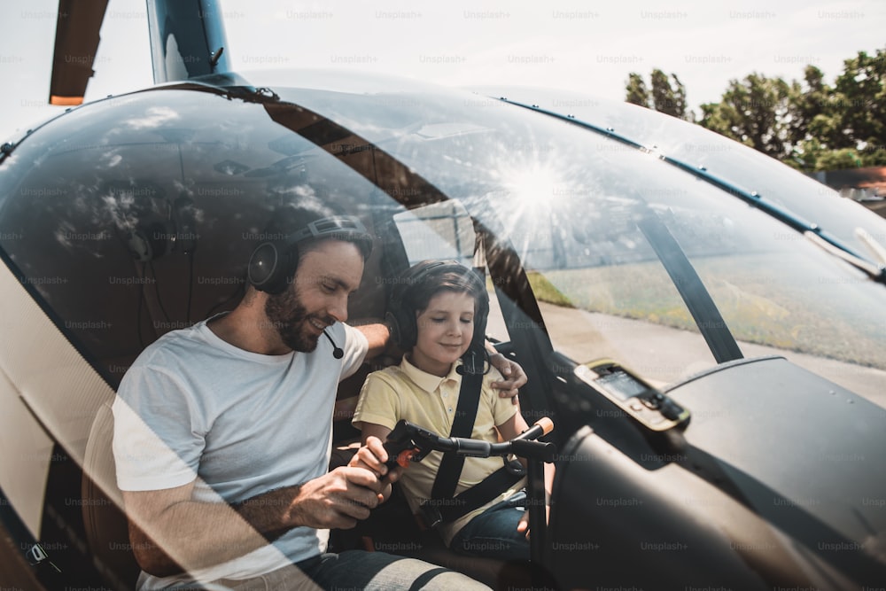 Beaming bearded dad teaching smiling kid to fly on rotorcraft. They locating inside of it while wearing headsets and communicating