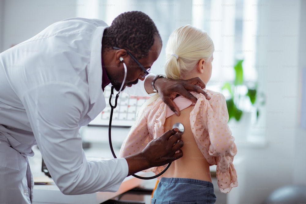 女の子を調べる。ブロンドの女の子を診察しながら聴診器を使用する浅黒い肌のプロの小児科医