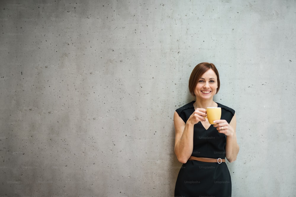 Un retrato de una joven y alegre mujer de negocios de pie en la oficina, sosteniendo una taza de café.