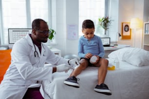 Poner yeso. Pediatra de piel oscura con bata blanca poniendo yeso en la pierna para un niño pequeño