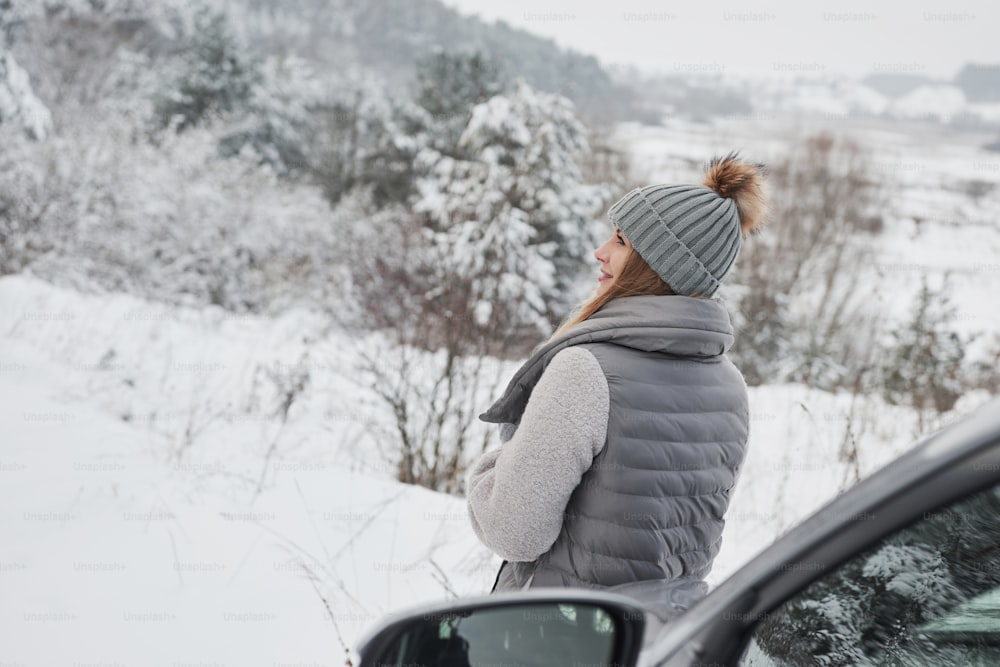 Looking at beautiful landscape. Pretty girl in warm clothes standing in the winter wood near the car.
