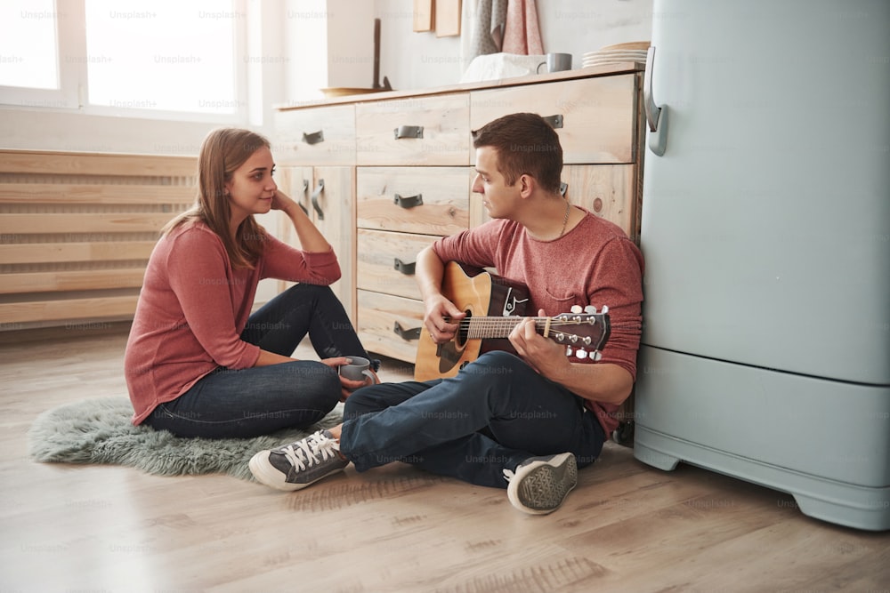 Sitting on the floor together and looking at each other. Young guitarist playing love song for his girlfriend in the kitchen.