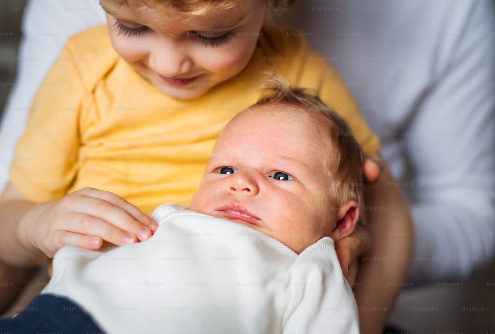A small toddler boy holding a newborn baby brother at home.
