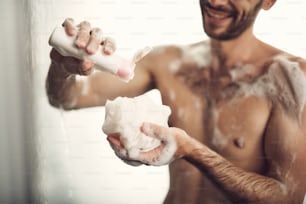 Close up photo of manly hands with gel and washcloth while standing under water in the bath room