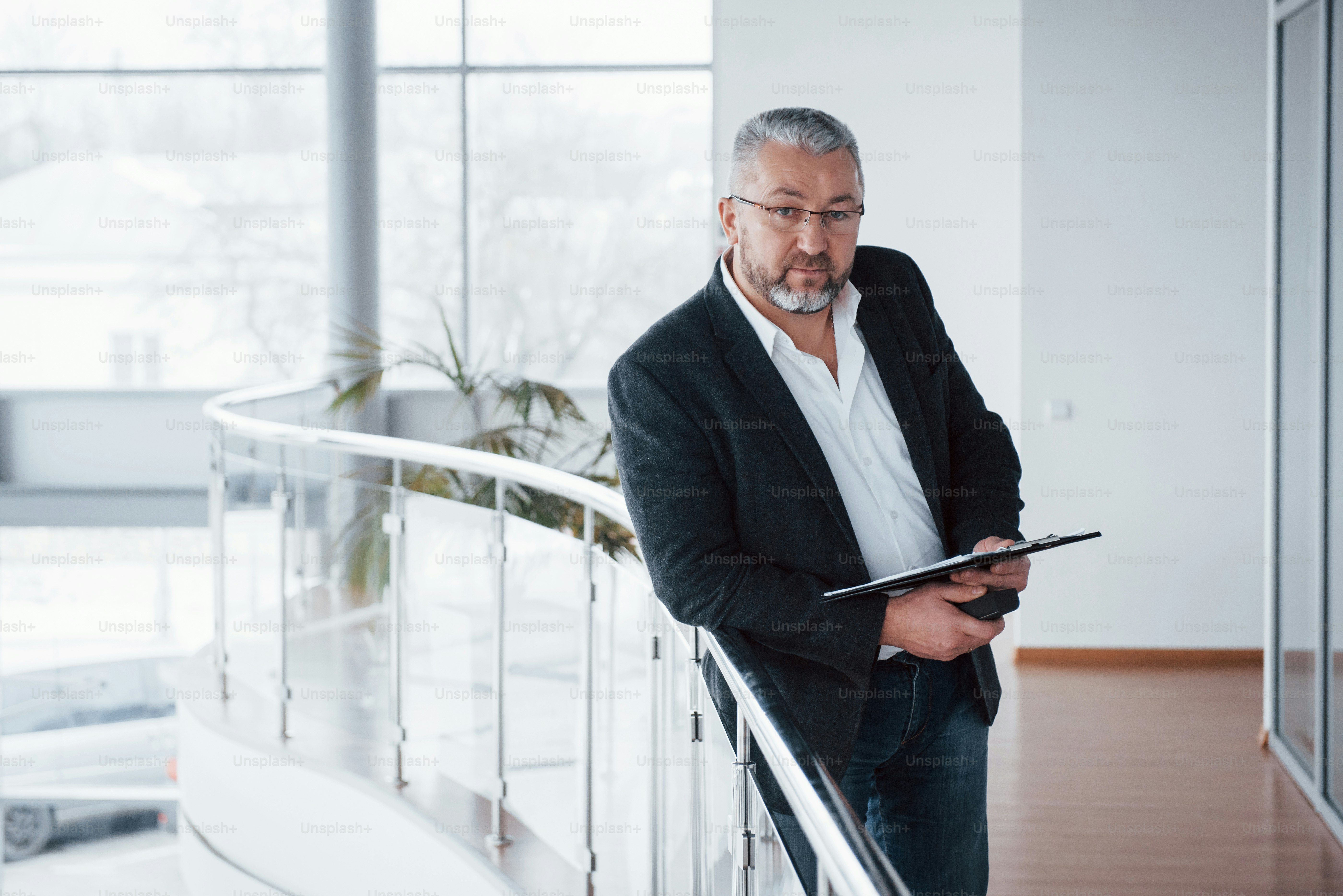 Good portrait. Photo of senior businessman in the spacious room with plants behind. Holding and reading documents