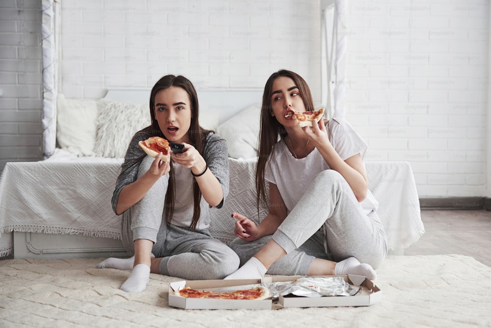 La comida siempre es más deliciosa cuando se ven películas. Hermanas comiendo pizza frente al televisor mientras se sientan en el suelo de una hermosa habitación durante el día.