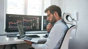 Anstrengender Tag. Professioneller männlicher Händler mit Brille, der den Finanzmarkt über einen Laptop analysiert, während er vor Computerbildschirmen mit Handelscharts im modernen Bürointerieur sitzt. Börse. Handelskonzept. Anlagekonzept