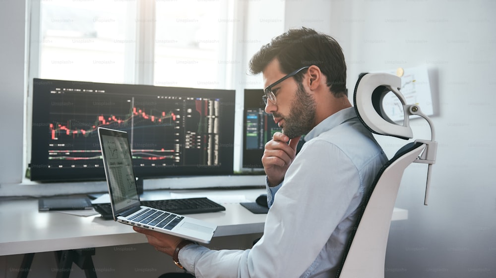 Busy day. Professional male trader wearing eyeglasses analyzing financial market via laptop while sitting in front of computer screens with trading charts in modern office interior. Stock exchange. Trade concept. Investment concept