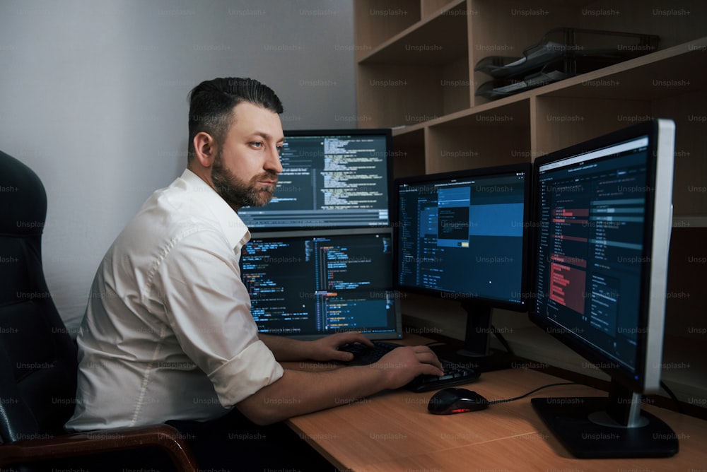Programmcodes sind überall. Der bärtige Mann im weißen Hemd arbeitet im Büro mit mehreren Computerbildschirmen in Indexcharts.