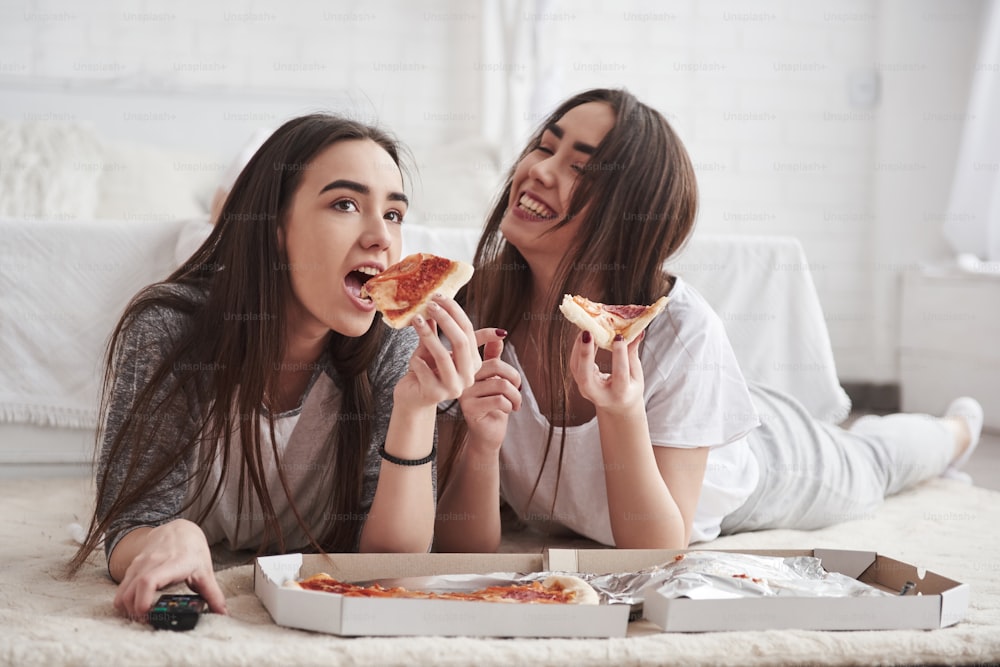 Semplicemente divertendosi. Sorelle che mangiano la pizza quando guardano la TV mentre sono sdraiate sul pavimento di una bella camera da letto durante il giorno.