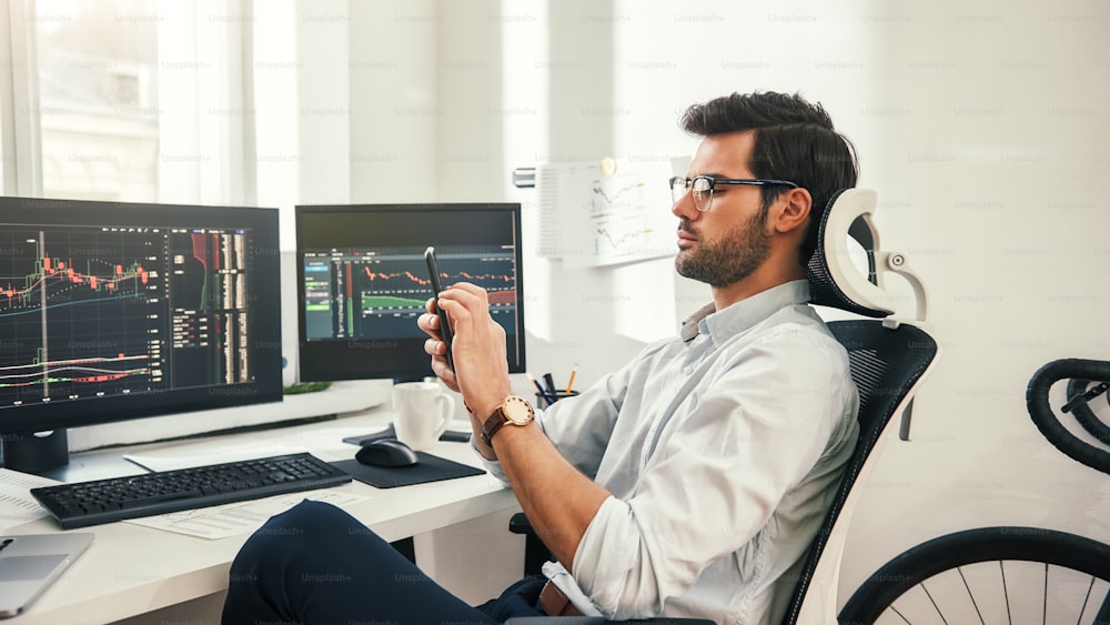 Trading online. Back view of young bearded trader in formal wear and eyeglasses using his smartphone while sitting in his office. Forex market. Trade concept. Investment concept