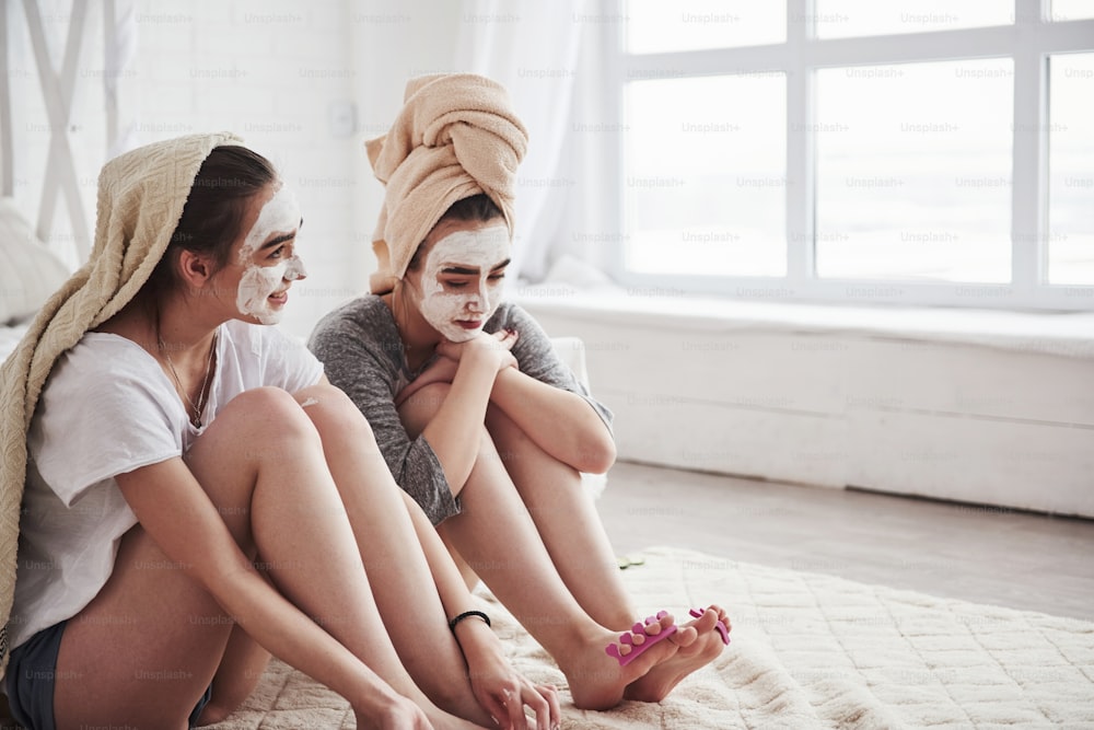 Two sisters twins in towels on heads and white beauty mask on the faces waiting for paint on the nails will become dry.