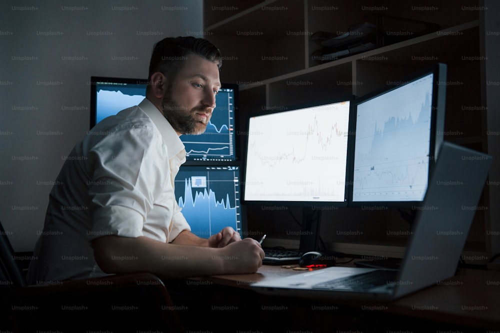 Looks at the laptop. Bearded man in white shirt works in the office with multiple computer screens in index charts.