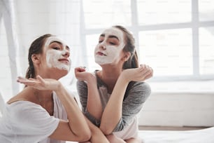 We are beautiful. Conception of skin care by using white mask on the face. Two female sisters have weekend at bedroom.