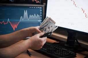 Vista de cerca de manos humanas sostiene dinero en la oficina con múltiples pantallas con gráficos e información diferente en ellas.
