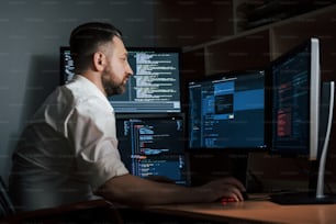 Programador adulto experiente. Homem barbudo de camisa branca trabalha no escritório com várias telas de computador em gráficos de índice.
