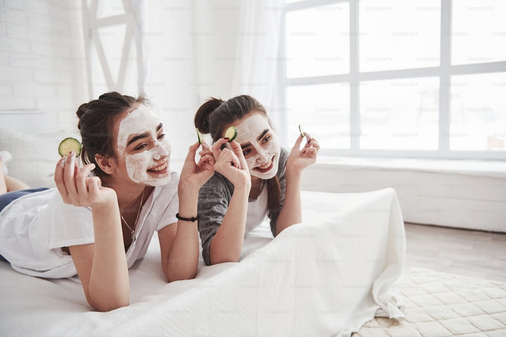 Posing for the cameramen. Conception of skin care by using white mask and cucumbers on the face. Two female sisters have weekend at bedroom.