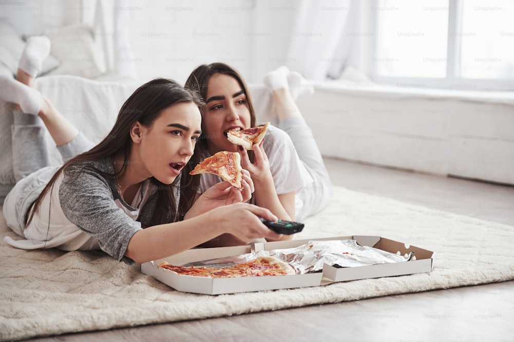 Freizeit. Schwestern essen Pizza, wenn sie fernsehen, während sie tagsüber auf dem Boden eines schönen Schlafzimmers liegen.