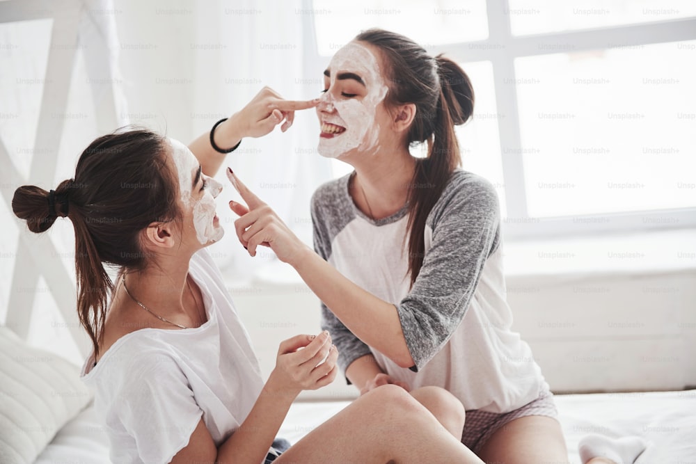Was zu tun ist, wenn Sie auf die Ergebnisse warten? Konzeption der Hautpflege mit weißer Maske im Gesicht. Zwei Schwestern haben Wochenende im Schlafzimmer.