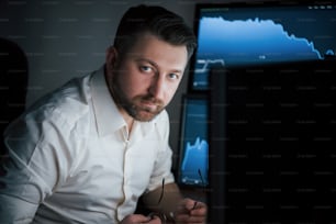 Looking into the camera. Bearded man in white shirt works in the office with multiple computer screens in index charts.