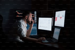 In dark night room. Bearded man in white shirt have a business call and works in the office with multiple computer screens in index charts.