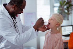 Open mouth wide. Dark-skinned pediatrician asking little girl with sore thorat to open mouth wide