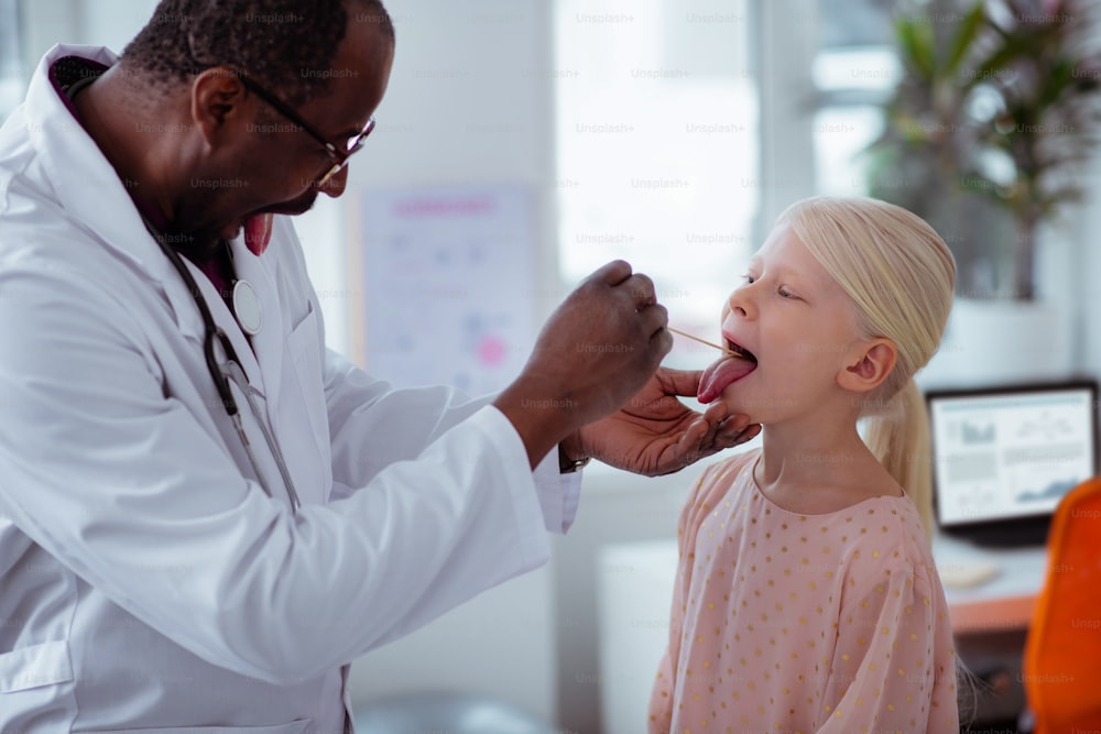 Abre bien la boca. Pediatra de piel oscura le pide a una niña con dolor de thorat que abra bien la boca