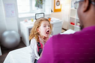 Showing throat. Blue-eyed curly girl opening mouth showing her throat while visiting doctor