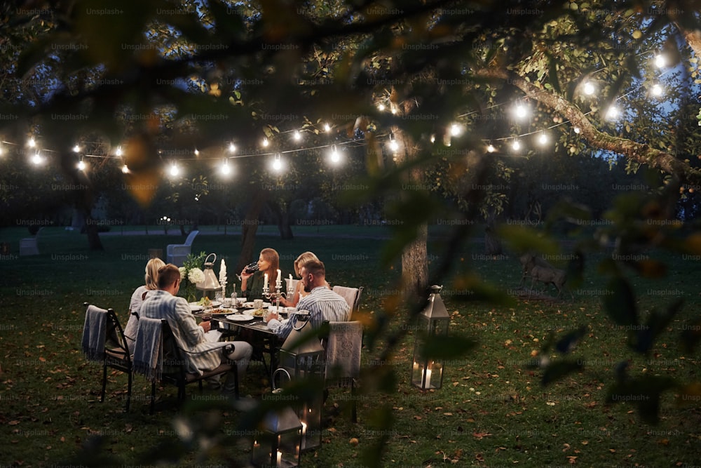 Foto através de galhos de árvores com folhas. À noite. Os amigos jantam no lindo lugar ao ar livre.