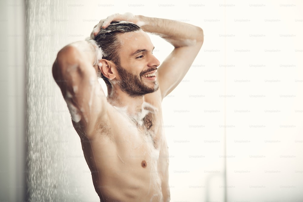 Un gars aux cheveux noirs mousse en train de prendre une douche. Homme souriant debout sous l’eau