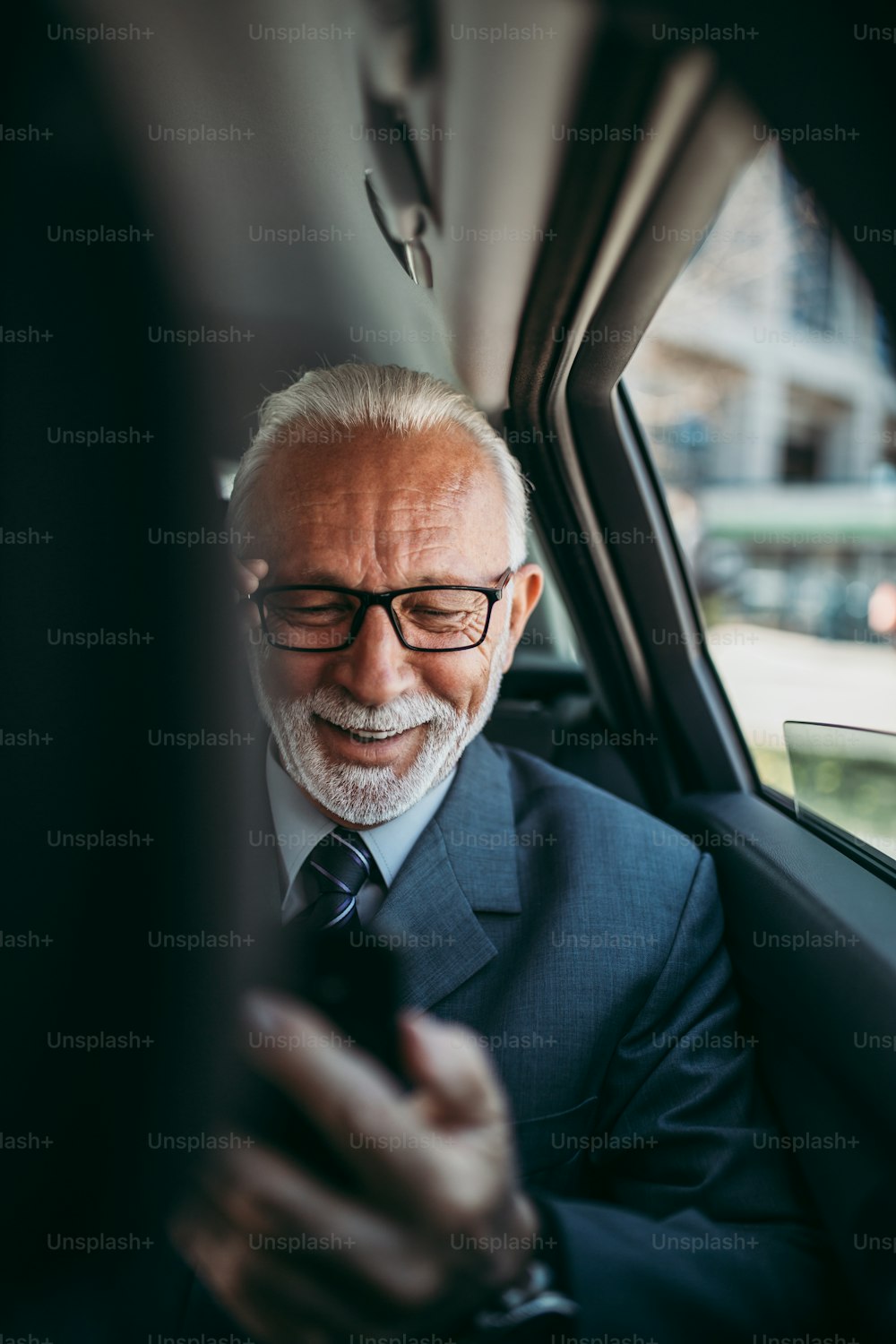 Good looking senior business man sitting on backseat in luxury car. He using his smart phone and reacting emotionally. Transportation in corporate business concept.