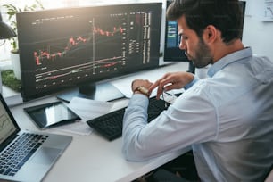 What time is it? Back view of bearded male stock market broker looking at watch on his hand while working with data and charts on computer screens in his modern office. Stock exchange. Trade concept. Investment concept