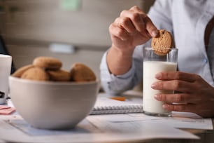 Femme d’affaires méconnaissable trempant un biscuit dans un verre de lait tout en prenant un petit-déjeuner au bureau.