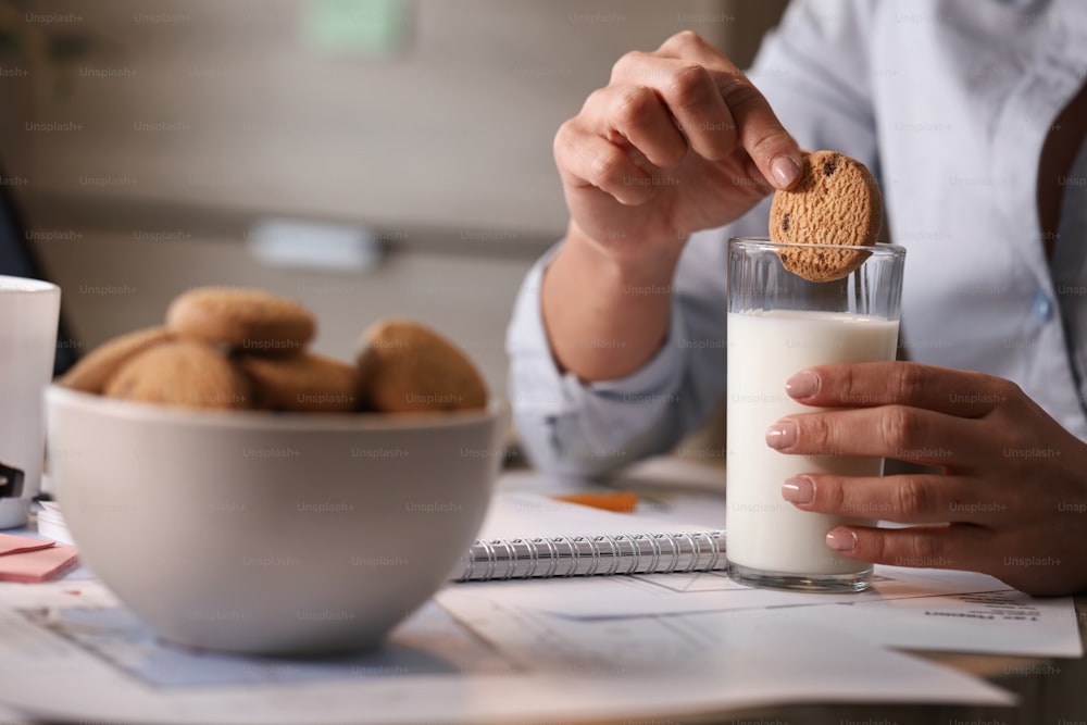 Unrecognizable businesswoman dipping cookie in glass of milk while having a breakfast in the office.
