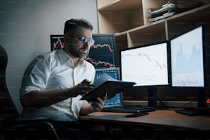 Konzentrierter ernsthafter erwachsener Unternehmer. Der bärtige Mann im weißen Hemd arbeitet im Büro mit mehreren Computerbildschirmen in Indexcharts.