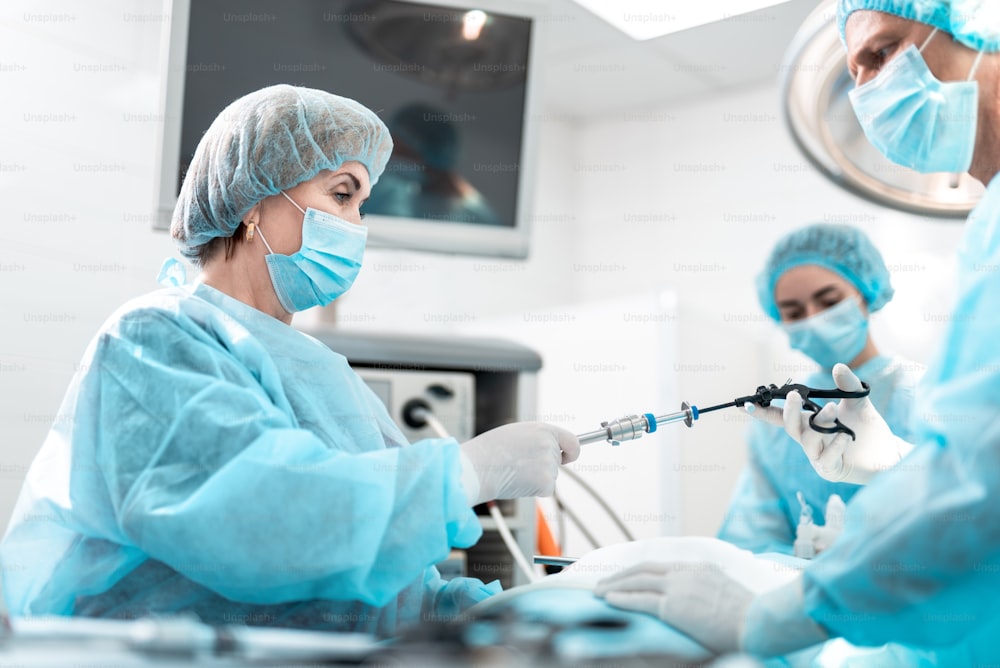 Side view portrait of female madical worker in sterile gloves helping doctor during surgical operation. They holding laparoscopic grasper with trocar