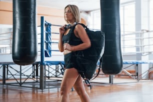 Walks to the garderobe. Adult female with black bag and headphones in the training gym.